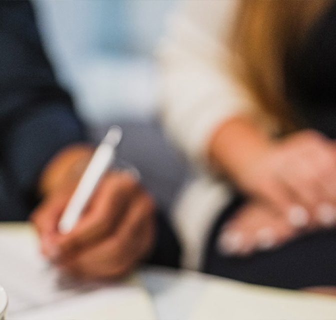 Two people signing documents at a table during a business meeting, one holding a white pen nearby.